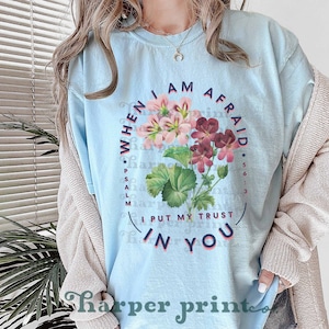 May include: Light blue t-shirt with a floral design and the text "WHEN I AM AFRAID *PSALM 56:3 I PUT MY TRUST IN YOU".