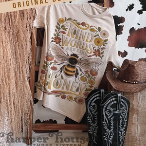 Peut inclure: Un t-shirt cr&egrave;me avec un motif floral et une abeille. Le texte dit "Kind Words are like Honey." Des bottes de cowboy noires et un chapeau marron sont &eacute;galement visibles. Le t-shirt est suspendu &agrave; une &eacute;chelle en bois.
