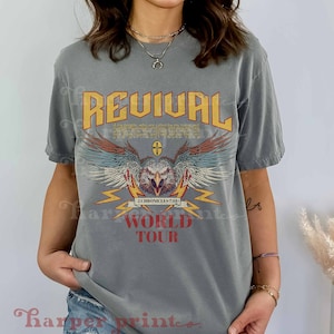 May include: A gray t-shirt with a vintage-style graphic design featuring an eagle with wings spread, lightning bolts, and the text "REVIVAL 2 CHRONICLES 7:14 WORLD TOUR".