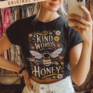 Peut inclure: T-shirt noir avec l'inscription "Kind Words Are Like Honey" et une illustration d'abeille. Le motif comprend des fleurs et des feuilles. Le t-shirt est port&eacute; avec un jean blanc.