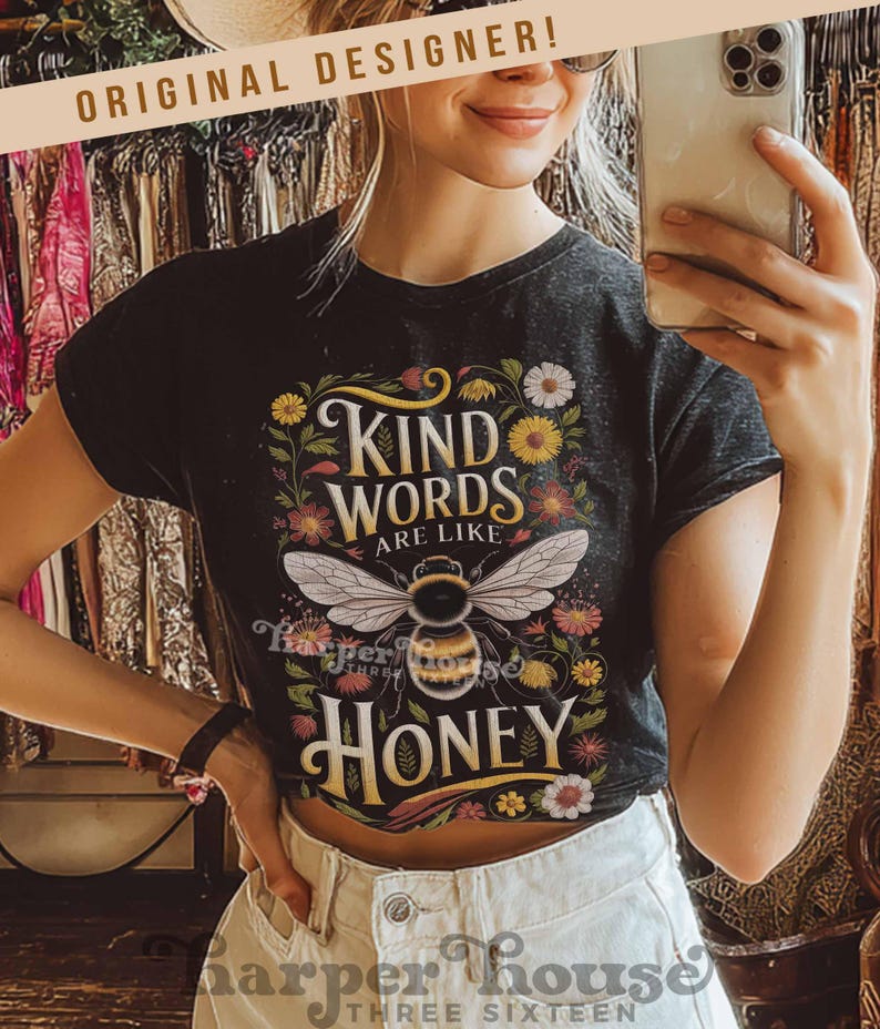 Peut inclure: T-shirt noir avec l'inscription "Kind Words Are Like Honey" et une illustration d'abeille. Le motif comprend des fleurs et des feuilles. Le t-shirt est port&eacute; avec un jean blanc.