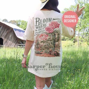 May include: A beige t-shirt with a vintage-style graphic of roses and a cactus. The graphic says "Bloom Where You Are Planted". The shirt also has the text "Harper House Three Sixteen" on the bottom.