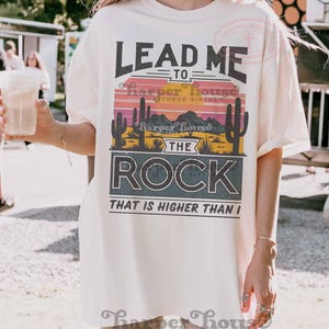 May include: A white t-shirt with a sunset graphic and the text "Lead me to the rock that is higher than I". The graphic features a desert landscape with cacti and mountains. The text "Harper House Three Sixteen" is also included.