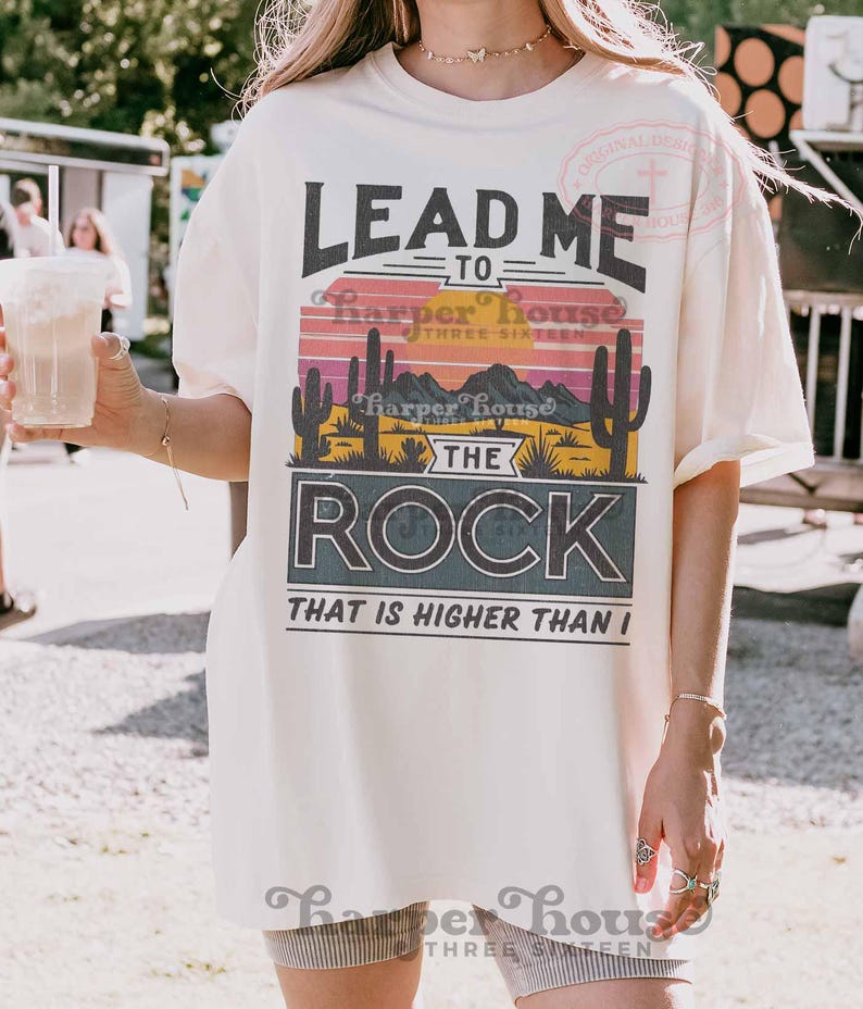 May include: A white t-shirt with a sunset graphic and the text "Lead me to the rock that is higher than I". The graphic features a desert landscape with cacti and mountains. The text "Harper House Three Sixteen" is also included.