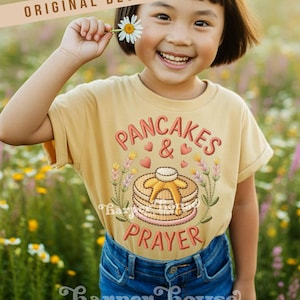 May include: A child wearing a mustard yellow t-shirt with the embroidered text "PANCAKES & PRAYER" and a stack of pancakes. The child is smiling and holding a daisy. The shirt also has the text "Harper House THREE SIXTEEN".
