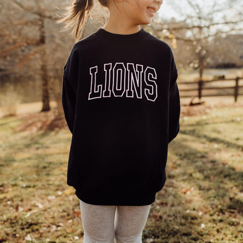 Puede incluir: Sudadera negra con el texto "LIONS" bordado en blanco en la parte delantera. La sudadera la lleva puesta un ni&ntilde;o.