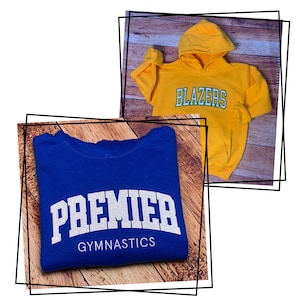 Puede incluir: Dos sudaderas con capucha. Una es azul con texto blanco que dice "PREMIER GYMNASTICS". La otra es amarilla con texto verde que dice "BLAZERS".