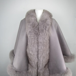 May include: A grey cape with a faux fur collar and trim. The cape is displayed on a mannequin and has a soft, flowing design. The fur trim is a lighter shade of grey, adding a luxurious touch to the outerwear.