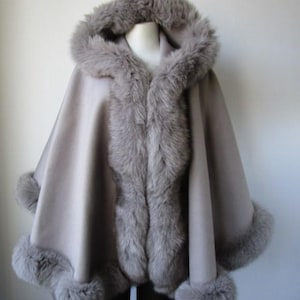 May include: A grey hooded cape with faux fur trim. The cape features a hood, a fur-lined front, and fur trim around the cuffs. The cape is a neutral colour, suitable for various outfits.
