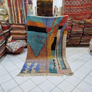 May include: A colorful, geometric patterned rug with a turquoise, orange, and black design. The rug has a fringed edge and is displayed in a shop with stacks of other rugs in the background.