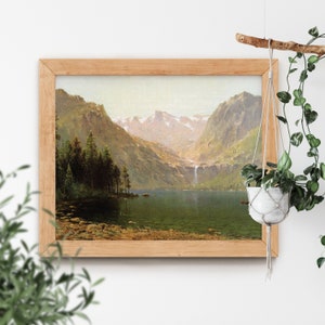 May include: A framed landscape painting depicts a serene mountain lake scene. The artwork features a waterfall, lush green trees, and snow-capped mountains under a soft, diffused light. The frame is made of light-colored wood.
