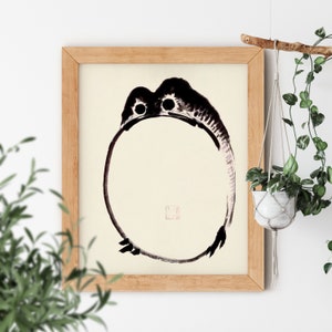 May include: A black and white ink painting of a frog in a circle, framed in a light wood frame.