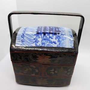 May include: A tiered black lacquer food carrier with a high arched handle. The top lid features a blue and white ceramic panel with a double happiness symbol. The black sides are decorated with intricate gold and red floral and leaf motifs.