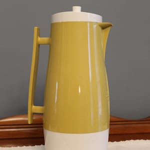 May include: A vintage yellow and white plastic pitcher with a handle and a lid. The pitcher has a tall, slender shape and a spout for pouring. The handle is a matching yellow color. The pitcher is likely from the mid-20th century.