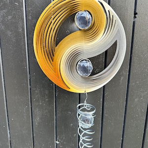 May include: A decorative wind spinner in a yin-yang design, featuring a gradient of gold to silver tones. Two clear crystal spheres are suspended from the spinner, with a spiral of crystals below. The background is a dark wooden surface.