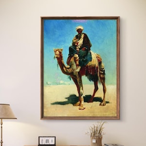 Puede incluir: Una pintura al óleo representa a una persona montada en un camello en un paisaje desértico. El jinete lleva un turbante y ropa oscura. El camello es de color canela con mantas de silla de montar coloridas. El fondo presenta un cielo azul y terreno arenoso, todo dentro de un marco de madera.