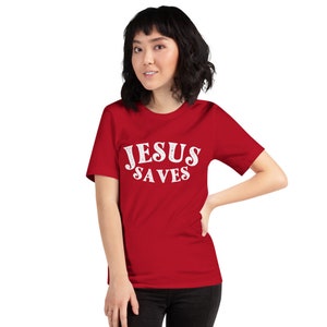 Pu&ograve; includere: Una maglietta rossa a maniche corte con il testo "JESUS SAVES" in lettere bianche.