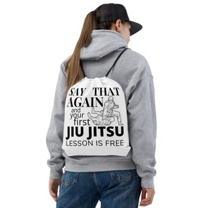 Op de afbeelding: Witte gymtas met zwarte tekst en illustraties. De tekst luidt: "SAY THAT AGAIN and your first JIU JITSU LESSON IS FREE." De illustratie toont twee Jiu Jitsu-beoefenaars. De tas heeft zwarte trekkoorden.