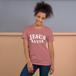 Pu&ograve; includere: Una donna indossa una maglietta rosa polverosa con il testo "JESUS SAVES" in lettere bianche.