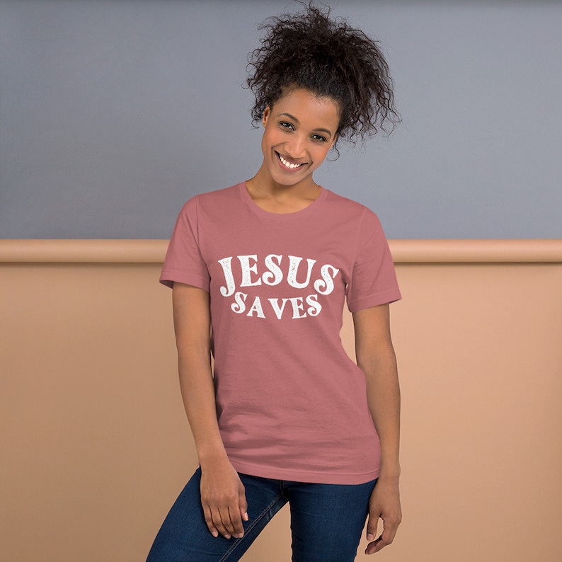 Pu&ograve; includere: Una donna indossa una maglietta rosa polverosa con il testo "JESUS SAVES" in lettere bianche.