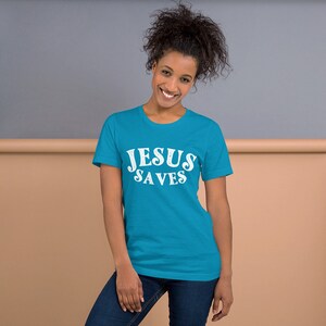 Pu&ograve; includere: Una maglietta blu turchese con il testo "JESUS SAVES" in bianco, lettere invecchiate.