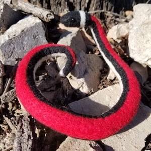 Puede incluir: Una serpiente de fieltro hecha a mano en posición enrollada. La serpiente tiene un cuerpo rojo con rayas negras y blancas. La serpiente descansa sobre un lecho de rocas y madera.
