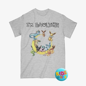 Puede incluir: Camiseta gris claro con la frase "I'M EEVOLVING" en letras negras. La camiseta tiene un gráfico de varias criaturas de dibujos animados dispuestas alrededor de una luna creciente amarilla. Una pegatina azul y amarilla "KIDS SIZE" está en la esquina inferior derecha.
