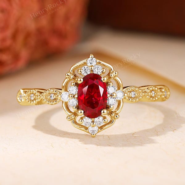 Puede incluir: Un anillo de oro con una piedra roja central de corte ovalado rodeada por un halo de pequeñas piedras blancas. El engaste tiene un marco de filigrana intrincado y la banda muestra un diseño texturizado a lo largo de su longitud.