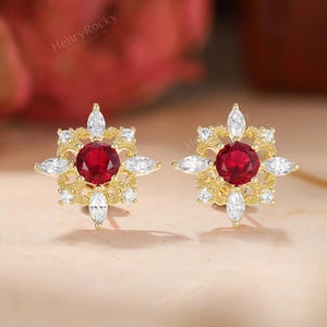 May include: A pair of gold stud earrings. Each earring features a central, round, deep red gemstone surrounded by smaller white gemstones and gold accents, creating a starburst design. The earrings are set against a soft, neutral background.