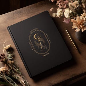 May include: A black journal with the words "SHADOW ORACLE" in gold lettering. The cover features a gold illustration of hands and a crescent moon. A gold pen and dried flowers are on a wooden surface.