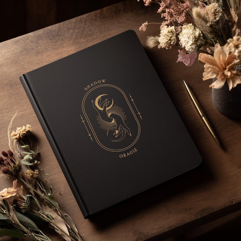 May include: A black journal with the words "SHADOW ORACLE" in gold lettering. The cover features a gold illustration of hands and a crescent moon. A gold pen and dried flowers are on a wooden surface.