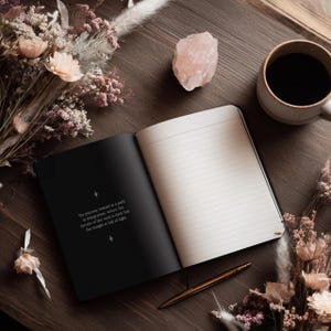 May include: An open black notebook with lined pages, a gold pen, and a cup of coffee on a wooden surface. A pink crystal and dried flowers are also present. The text on the left page reads: "The journey inward is a path to integration, where the terrain of the void is dark but the insight is full of light."