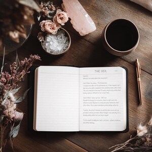 May include: An open journal with lined pages and the title "THE VEIL" sits on a wooden surface, surrounded by a pen, a cup, crystals, and dried flowers. The journal's pages are filled with handwritten text.