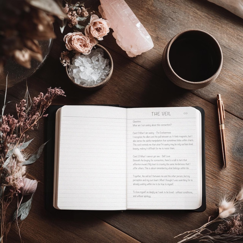 May include: An open journal with lined pages and the title "THE VEIL" sits on a wooden surface, surrounded by a pen, a cup, crystals, and dried flowers. The journal's pages are filled with handwritten text.