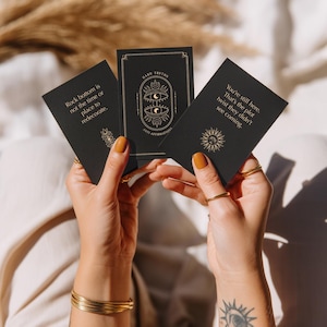 Può includere: Tre carte nere con testo e illustrazioni dorate sono tenute davanti a uno sfondo morbido. Le carte recitano "Rock bottom is not the time or place to redecorate", "HARD TRUTHS ANTI-AFFIRMATIONS" e "You're still here. That's the plot twist they didn't see coming."
