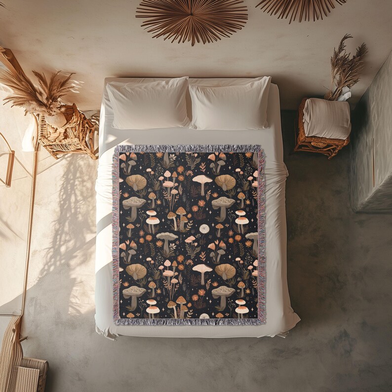 May include: A dark-colored throw blanket with a repeating pattern of mushrooms, flowers, and foliage. The blanket has a fringed edge and is placed on a white bed. The color palette includes shades of brown, beige, and orange.