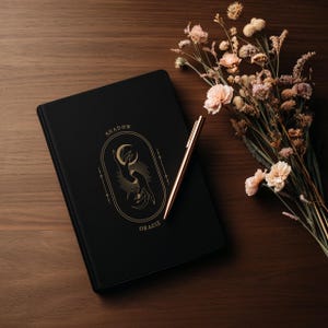 May include: A black journal with gold lettering that reads "SHADOW ORACLE" and a gold pen rest on a wooden surface. A bouquet of dried flowers with pink and cream-colored blooms sits to the right of the journal.
