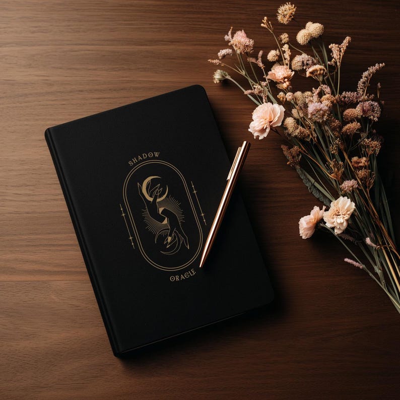May include: A black journal with gold lettering that reads "SHADOW ORACLE" and a gold pen rest on a wooden surface. A bouquet of dried flowers with pink and cream-colored blooms sits to the right of the journal.