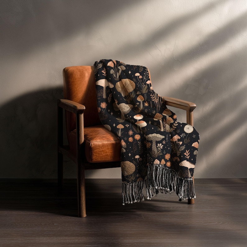 May include: A black throw blanket with a repeating pattern of mushrooms, plants, and flowers in shades of brown, tan, and orange. The blanket is draped over a brown leather armchair and has a fringe along the bottom edge.