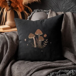 May include: Black square throw pillow featuring a mushroom illustration. The design includes three brown mushrooms with white spots, along with sprigs of grass, leaves, and small star accents. The pillow is placed on a gray blanket.