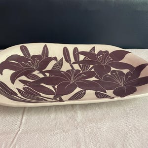May include: A cream-colored, oblong ceramic serving platter with a floral design. The flowers are a deep purple color and are stylized lilies. The platter is on a white surface with a dark blue background.