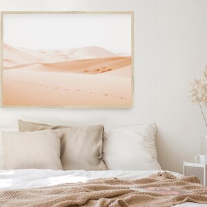 May include: A framed photograph of a desert landscape with footprints in the sand. The photo is in a light wood frame and is hanging on a white wall above a bed with beige bedding.