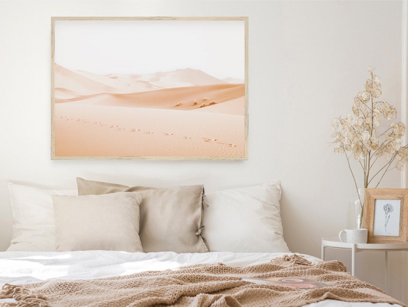 May include: A framed photograph of a desert landscape with footprints in the sand. The photo is in a light wood frame and is hanging on a white wall above a bed with beige bedding.