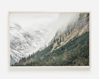 May include: A misty mountain landscape with snow-capped peaks and a lush green forest in the foreground. The mountains are covered in fog, creating a dramatic and ethereal atmosphere.