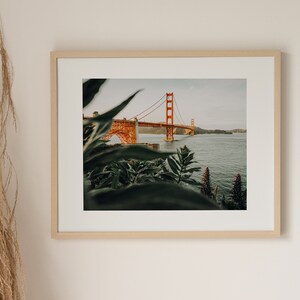 May include: Framed print of the Golden Gate Bridge in San Francisco. The orange bridge is set against a backdrop of water and sky, with green plants in the foreground. The print is presented in a light wooden frame, enhancing the natural aesthetic.