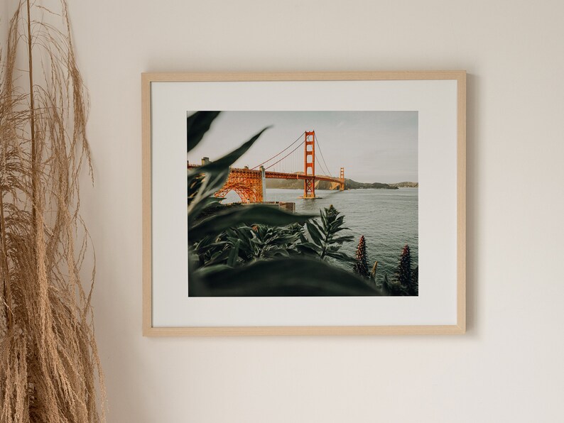 May include: Framed print of the Golden Gate Bridge in San Francisco. The orange bridge is set against a backdrop of water and sky, with green plants in the foreground. The print is presented in a light wooden frame, enhancing the natural aesthetic.