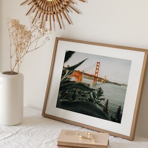 May include: A framed print of the Golden Gate Bridge in San Francisco, California, with an orange-red paint color. The print is in a light brown wooden frame, and is on a white surface. A white vase with dried flowers and a decorative candle holder are also present.