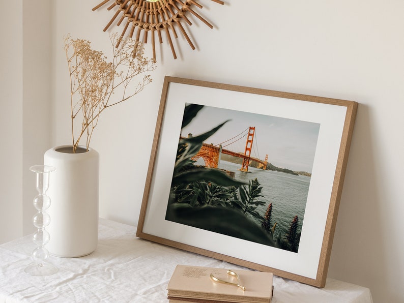 May include: A framed print of the Golden Gate Bridge in San Francisco, California, with an orange-red paint color. The print is in a light brown wooden frame, and is on a white surface. A white vase with dried flowers and a decorative candle holder are also present.