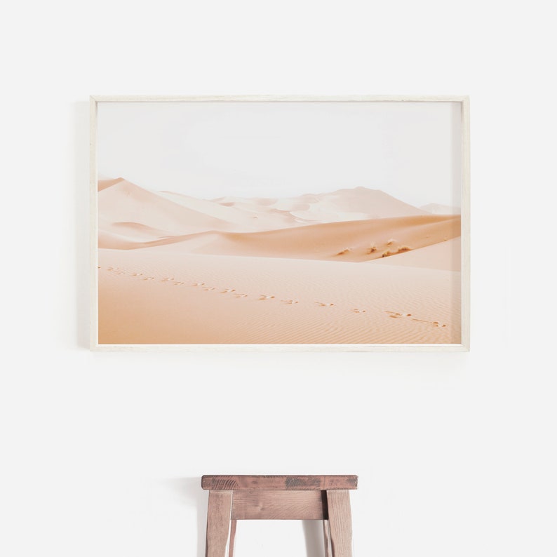 May include: A framed photograph of a desert landscape with sand dunes and footprints in the sand. The photograph is in a white frame and is hanging on a white wall.