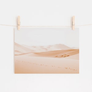 May include: A photograph of a desert landscape with sand dunes and footprints in the sand. The sky is a pale blue and the sand is a warm beige color.